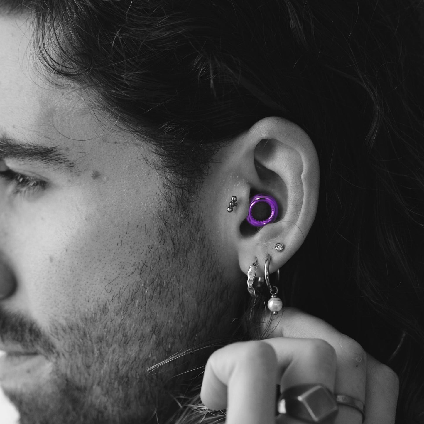 צילום תקריב בשחור-לבן של פרופיל פניו של גבר עם שיער כהה ארוך וגלי וזקן קצר. הוא עונד מספר פירסינגים באוזן: שלושה עגילים כדוריים קטנים, עגיל צמוד כסוף עם אבן קטנה, שני עגילי חישוק בתנוך האוזן (אחד מהם עם פנינה תלויה), ואטמי אוזניים איכותיים סגולים בחלק הפנימי של האוזן, שהם האלמנט הצבעוני היחיד בתמונה. ידו של הגבר נראית בחלק התחתון של התמונה, עונדת טבעת גדולה